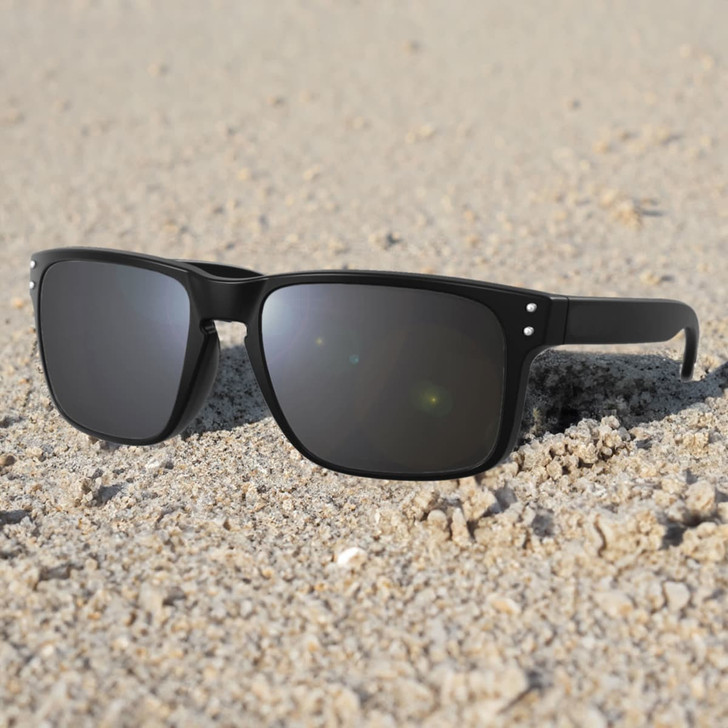 A pair of black sunglasses resting on sandy ground, featuring a simple design and grey lenses.