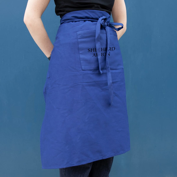 A blue long waist apron with a front pocket and tied at the waist, displayed against a blue background.
