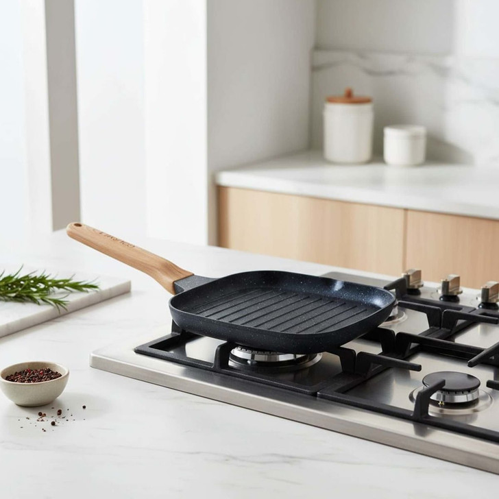 A square grill pan with a wooden handle, featuring a non-stick black surface, sits on a modern gas stovetop.