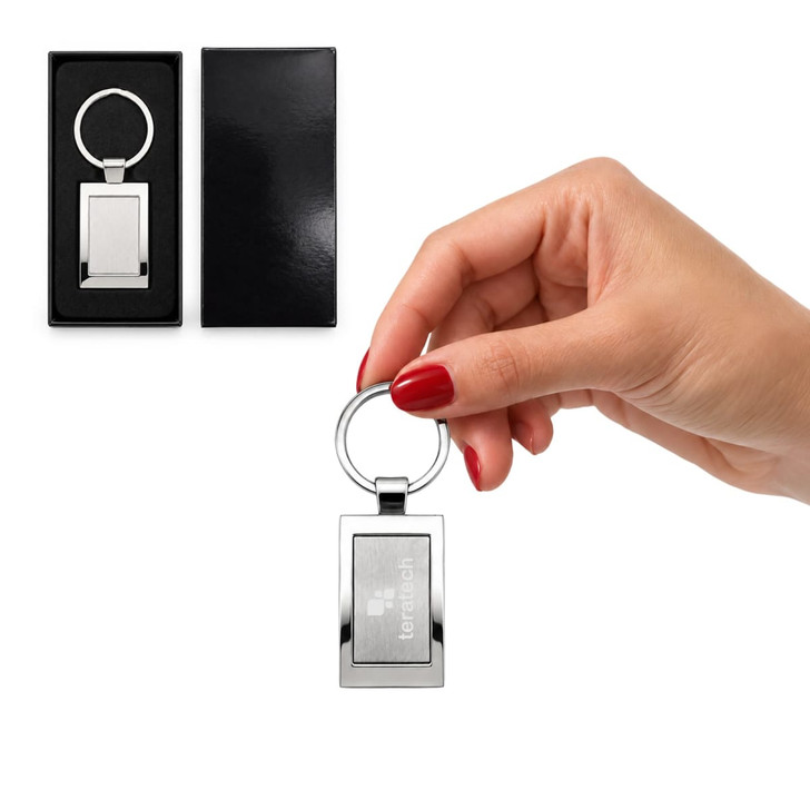A silver rectangular keytag held in a hand, displayed in a black gift box. The keytag features a logo.