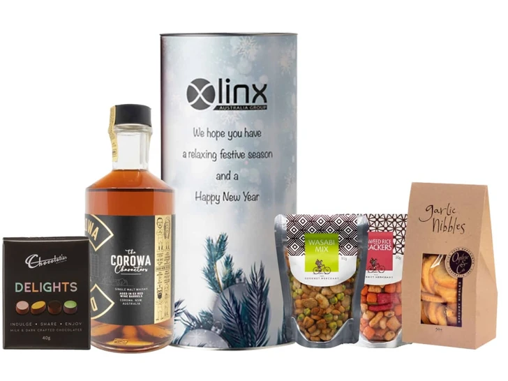 A whisky bottle, gourmet snacks, and chocolates arranged in a festive hamper with a cylindrical container.