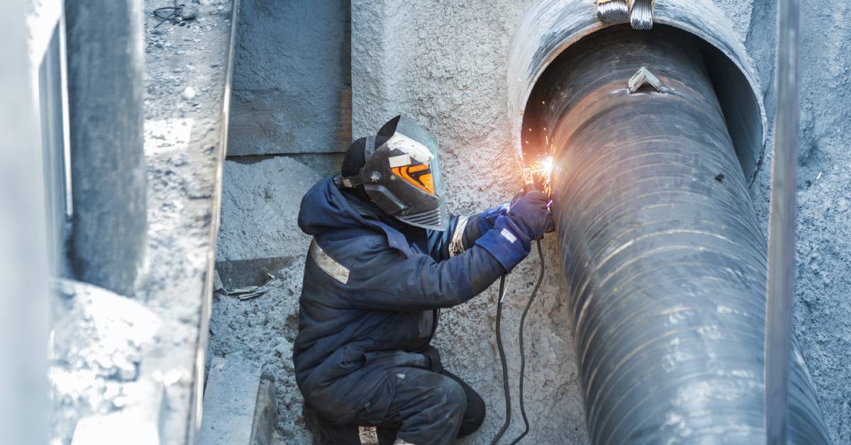 A person wearing protective clothing and a large mask welds a large steel pipeline coming out of a concrete fixture.