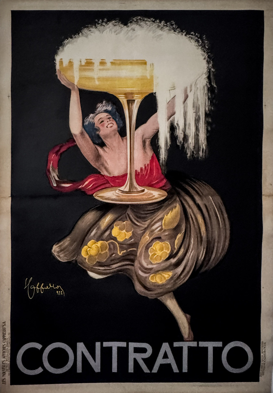 Asti Contratto 2 sheet by Leonetto Cappiello depicts a woman holding up large glass of bubbling champagne. The champagne is overflowing over the glass in a majestic shower.  She is wearing a red sash and flowing brown dress with accents of gold in the shape of grape vines.