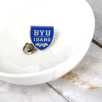 BYU Idaho Pin, blue and silver shield-shaped pin with white text and floral design, displayed on a white dish, university logo pin.