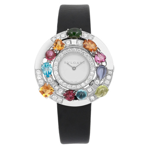 An image of a pre-owned wristwatch taken from a front full frontal view. The watch features a round face with a silver-tone bezel adorned with a variety of colorful gemstones and small diamonds. The dial is simple with two hands and the brand name visible. It has a black strap that extends vertically from the top and bottom of the watch face.