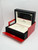 An image of an unworn wristwatch box. The box is open, showing a cream interior with the brand name "Tudor" visible. The exterior is black with a red bottom flap, positioned at a slight angle and captured from a close distance. The watch itself is not present in the image.
