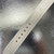 An image of a pre-owned Hermès women's wristwatch strap, viewed from a close-up angle. The strap is laid flat on a reflective surface, highlighting its light color and stitching details. The image captures the strap's adjustment holes and texture.