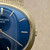 An image of a pre-owned Patek Philippe wristwatch, captured in a close-up view focusing on the right side of the watch face. The angle highlights the gold-toned bezel and crown against a deep blue dial with minimalistic gold hour markers. The watch is positioned slightly tilted, showing the textured band at the top edge.