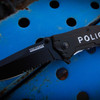 Close-up of the Tac-Force TF-525PD Police rescue folding knife resting on blue industrial steel, highlighting the assisted opening blade, half-serrated edge, and black aluminum handle with POLICE graphics.
