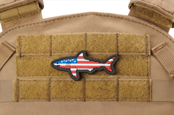 PVC morale patch shaped like a shark featuring a USA flag design, mounted on a tactical vest with hook-and-loop backing.