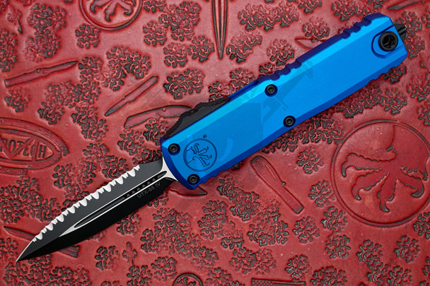 Microtech UTX-85 GEN IV automatic knife with blue aluminum handle and black full serrated double edge blade opened on red textured background.