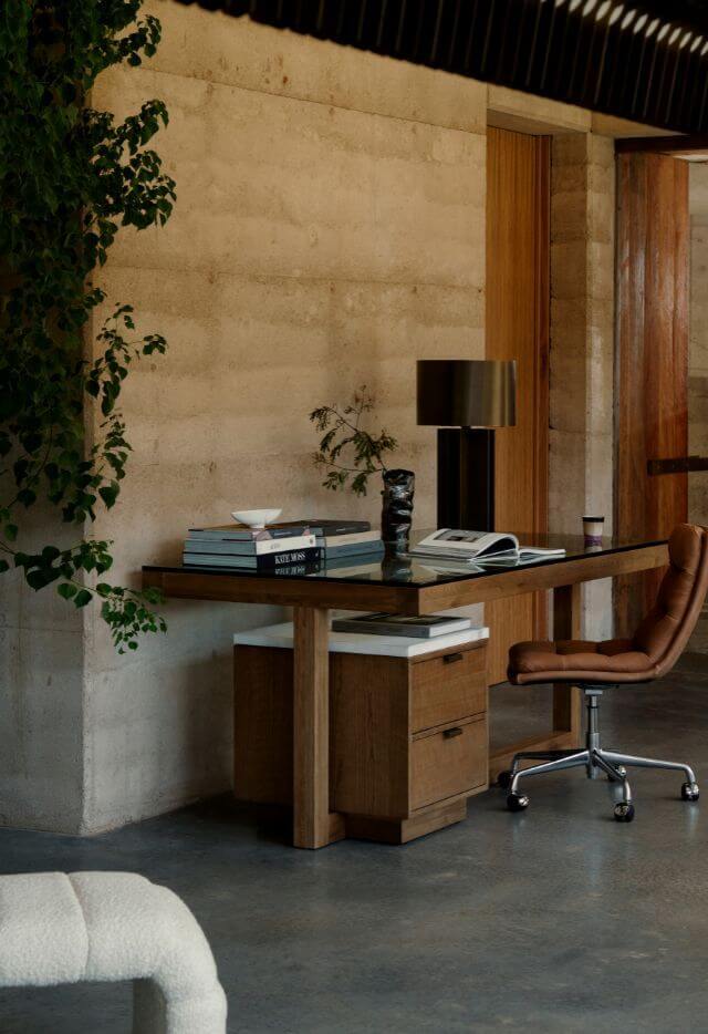 A modern office with a wooden desk featuring a glass top and marble drawers. The desk holds a stack of books, a brass lamp, and a small vase with a branch. A brown leather office chair with chrome wheels is pulled up to the desk. The room has a textured concrete wall and a leafy green plant hangs down the left side.