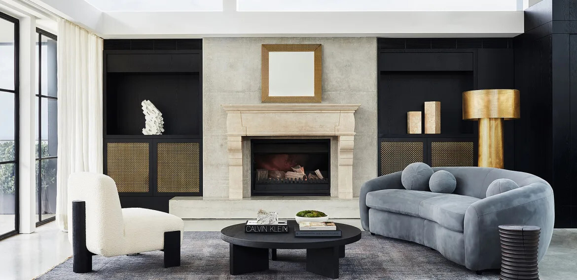 Atelier sofa in dusty blue curved velvet, styled beside a stone fireplace in a moody black-toned living room.