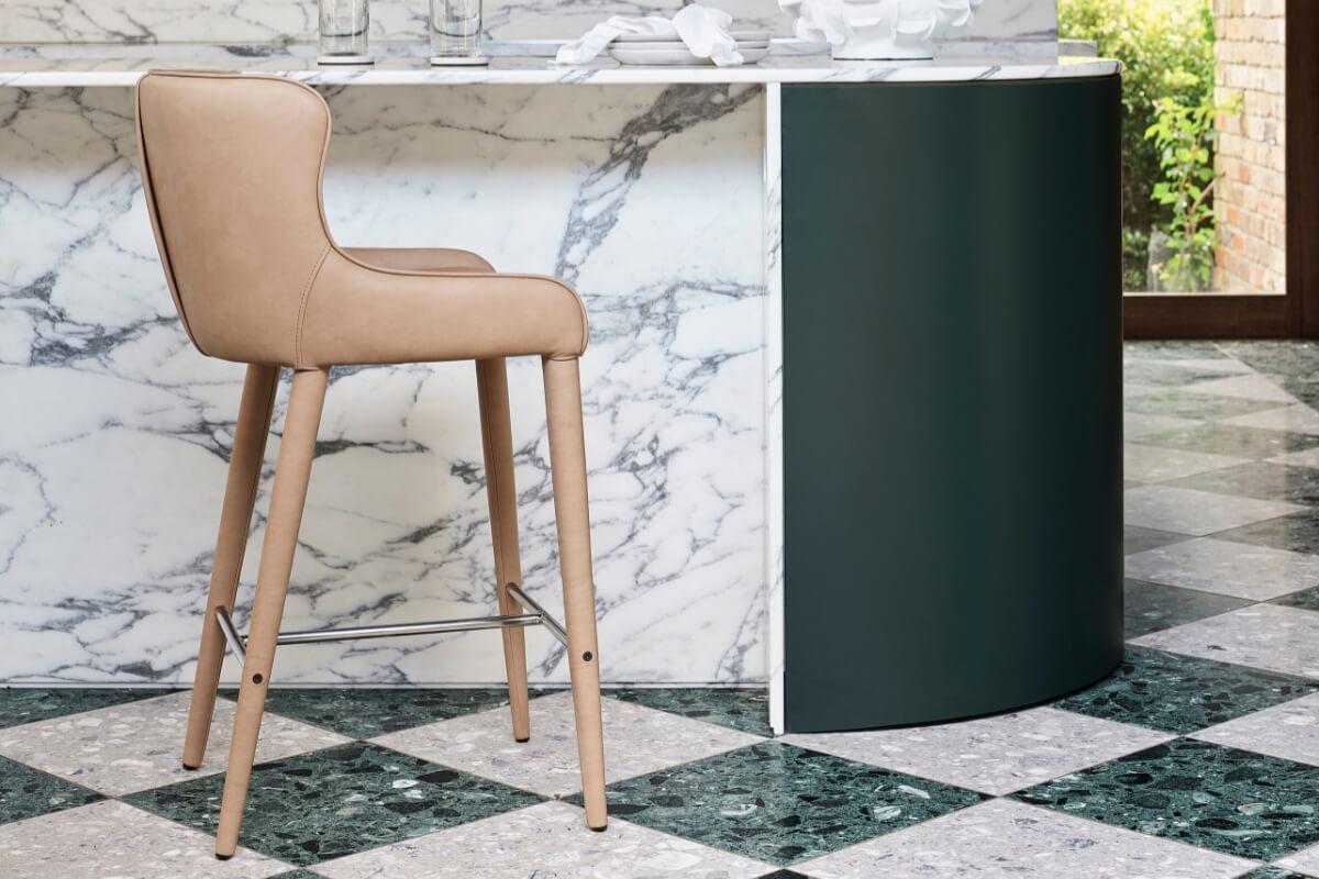 A Markson Counter Stool with a cushioned seat and gently rounded back is positioned at a kitchen counter with a white marble top and a dark green base. The kitchen floor is made of large, black and white checked tiles.