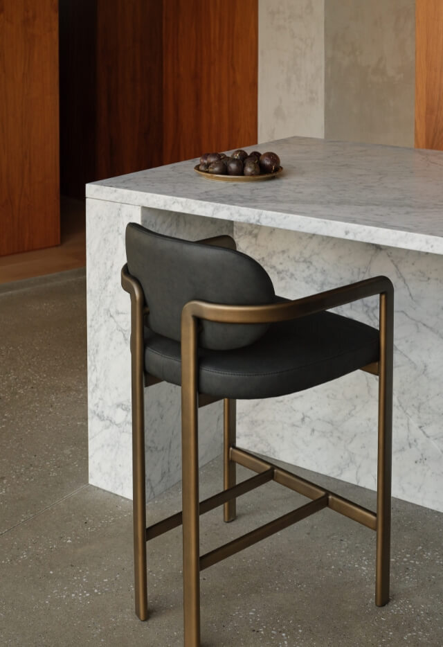A Stuttgart Counter Stool with a sculptural frame and a contoured, dark seat is positioned next to a white marble kitchen counter. A small plate of figs sits on the counter.