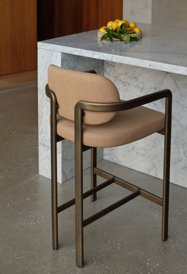 A Stuttgart Counter Stool with a sculptural frame and a contoured, light brown seat is positioned next to a white marble kitchen counter. A wooden bowl of lemons sits on the counter.