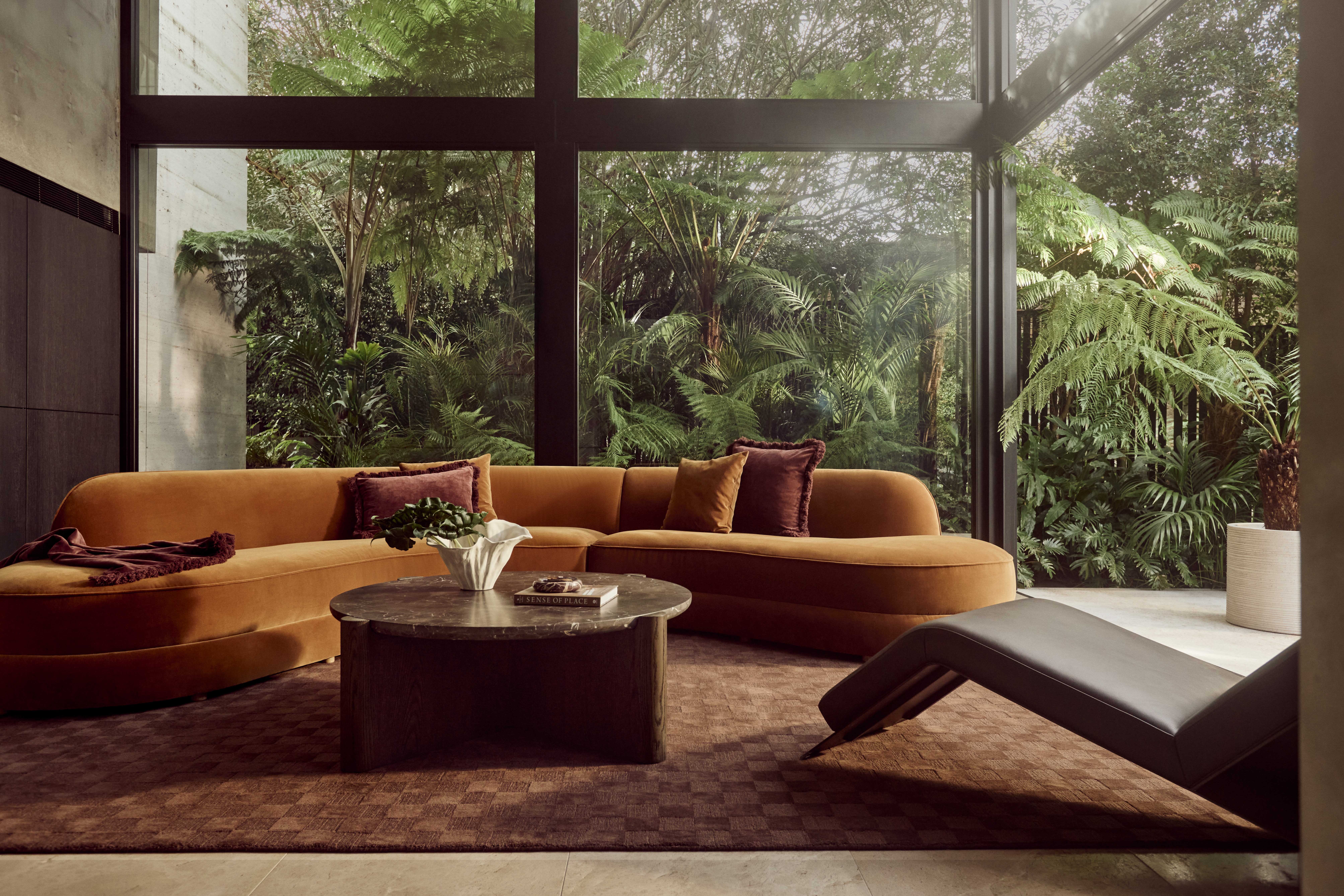 Coco Republic Margaux Curved Modular Sofa in mustard velvet, styled in a glass-walled living room with lush tropical greenery and a round stone coffee table.