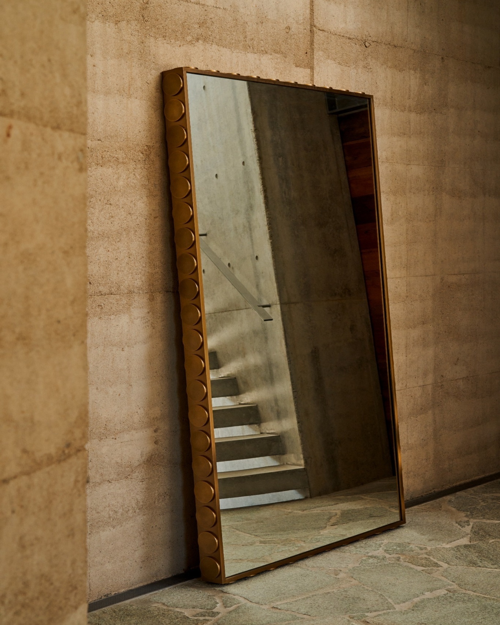 Outdoor mirror with stainless steel frame, leaning against a stone wall in an outdoor entryway.