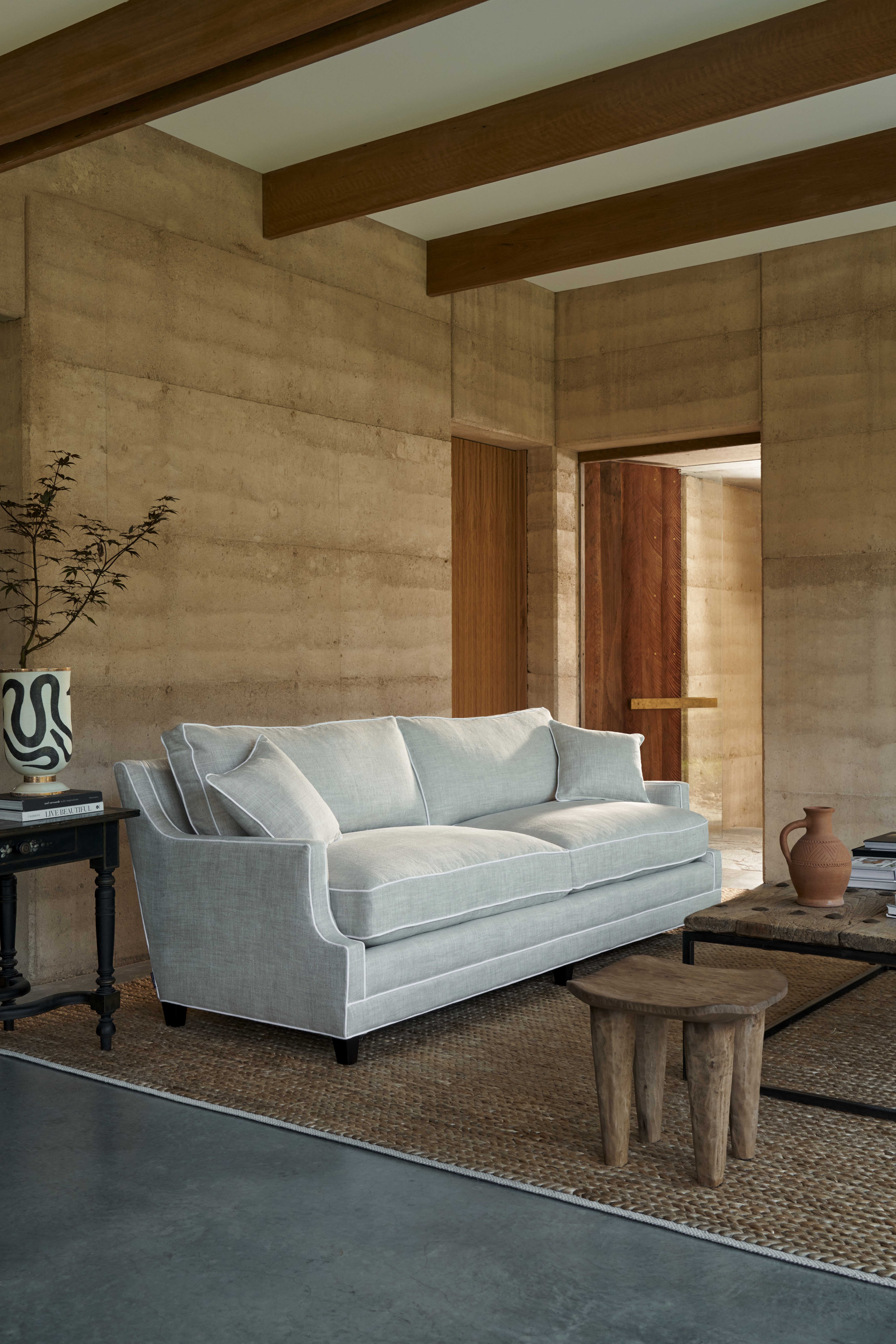 Coco Republic Ashley Sofa in soft blue linen, styled in a rammed-earth interior with rustic timber furniture and natural light.