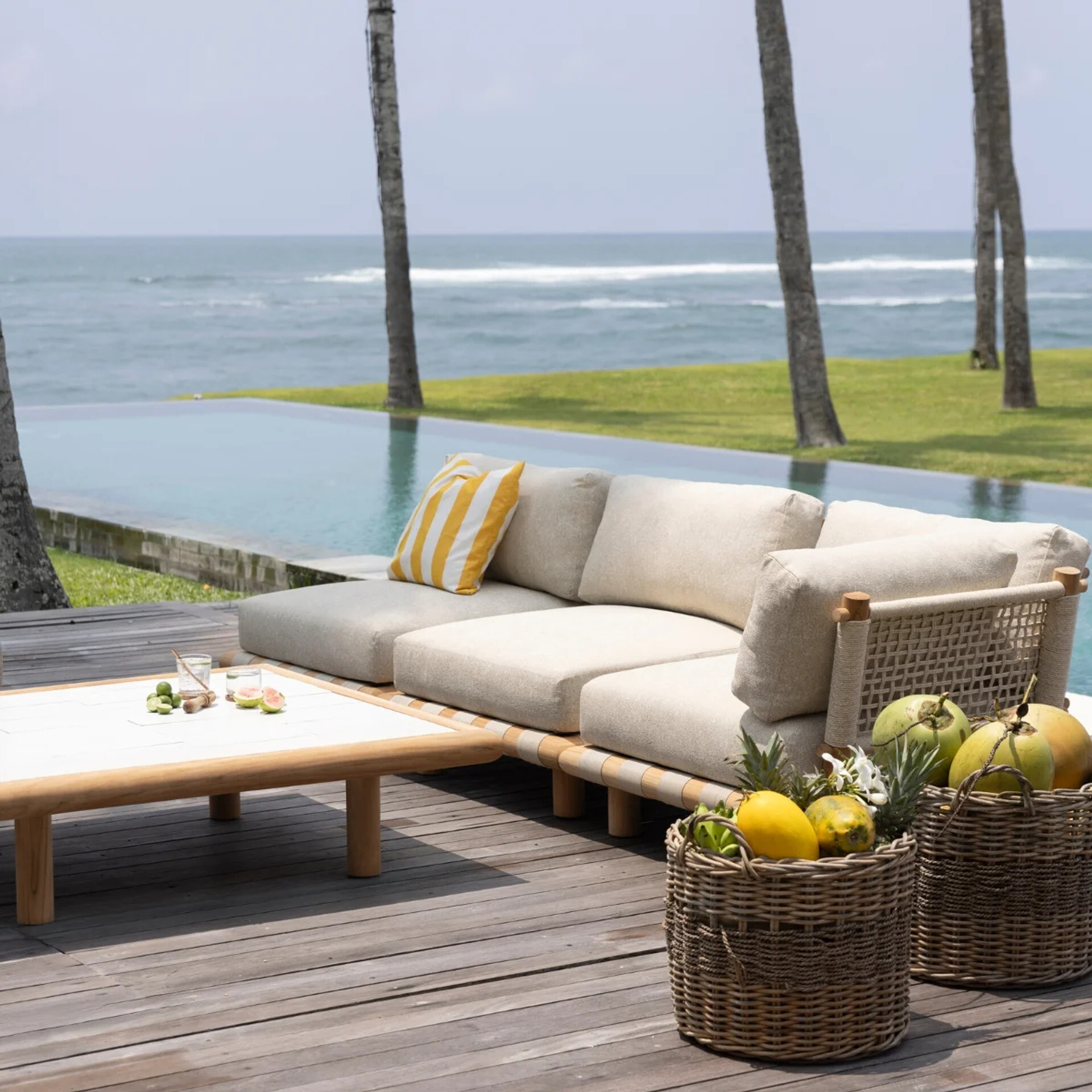 Coco Republic Tulum Outdoor Modular Sofa with teak frame and boucle cushions, styled poolside overlooking the ocean with woven baskets of tropical fruit.