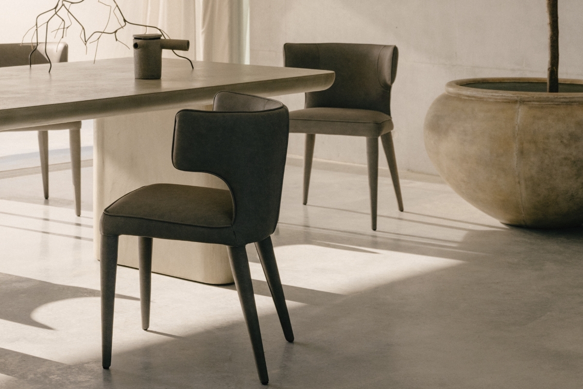 Coastal dining space featuring Melrose Dining Chairs in dark upholstery with curved backs, styled around a sculptural stone dining table overlooking the ocean.