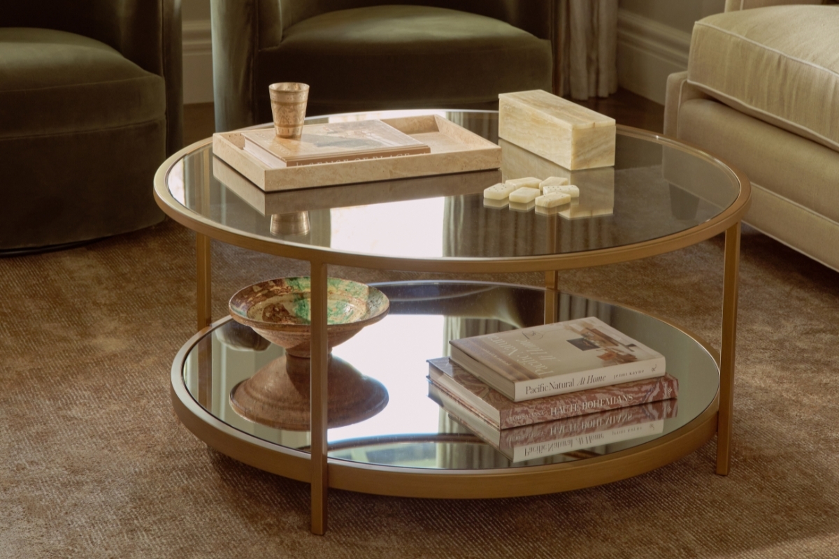 Gold-framed glass coffee table styled with books and decor, flanked by velvet armchairs in a soft-toned living room