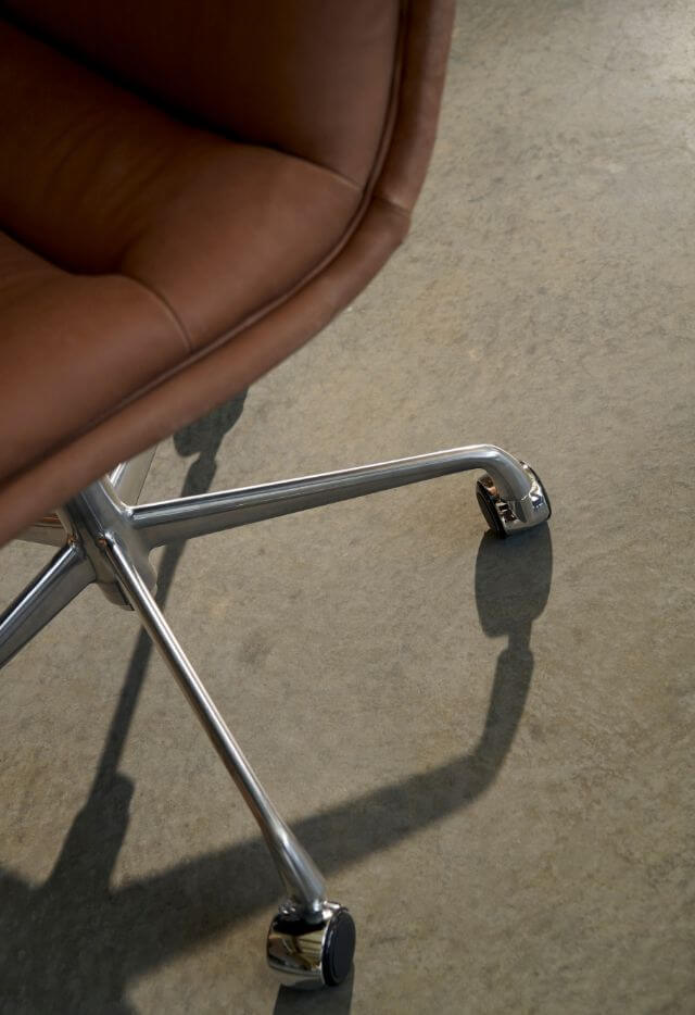 A close-up, low-angle shot of the chrome base and wheels of a brown leather office chair, casting a shadow on a concrete floor.