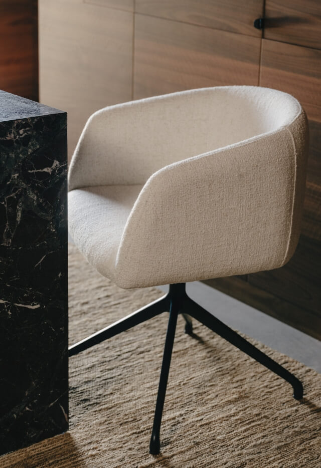 A cream-coloured upholstered desk chair with a brass metal four-point base sits on a textured beige rug next to a black marble desk.