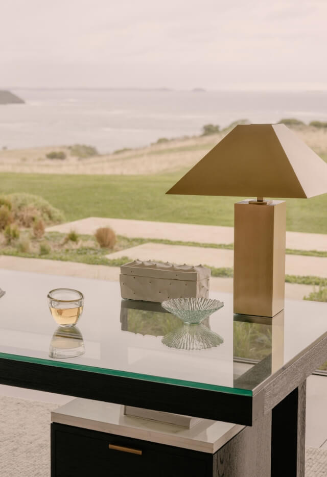 A modern desk with a gold lamp, a small gray box, a glass bowl, and a drink in a unique glass sits in front of a large window with a view of a grassy hill and the ocean.
