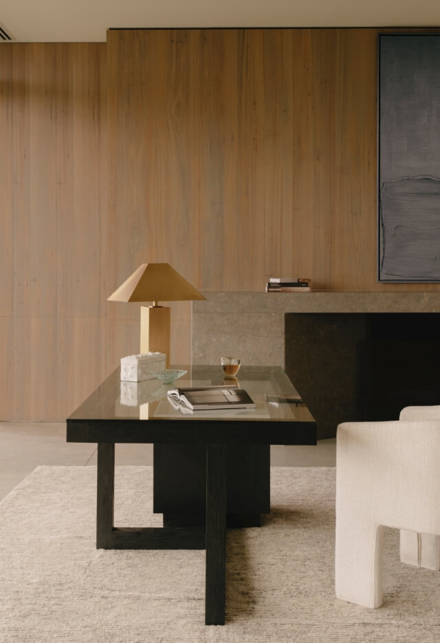 A close-up of a modern desk with a gold lamp, a grey box, an open book, and a cup on it. The desk has a glass top and a dark wooden base. In the background is a wood-panelled wall and a white upholstered chair is partially visible on the right.
