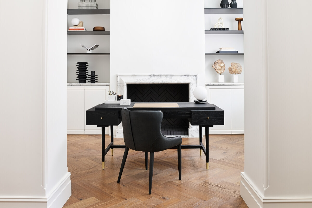 A stylish home office with a black wooden desk and matching black chair with brass accents. The desk is centered in the room in front of a white fireplace with a black patterned hearth. Built-in shelves on either side display various decorative objects.