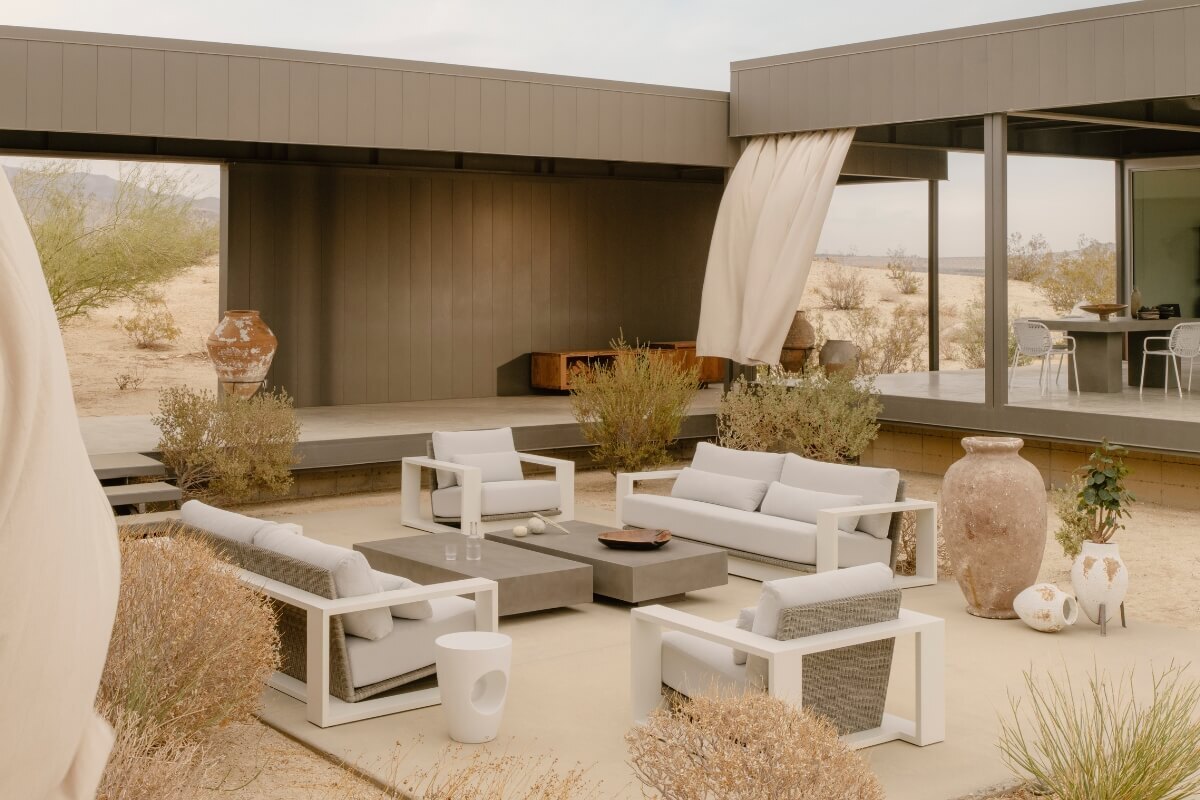Modern outdoor lounge setting with white frame armchairs and grey cushions, surrounding a low rectangular concrete coffee table.
