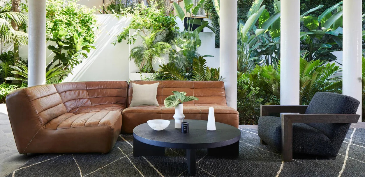 Shabby Sectional Sofa by Timothy Oulton in tan leather, styled on a covered outdoor terrace surrounded by greenery.