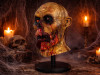 A grotesque zombie mask with decaying skin, blood, and exposed teeth is displayed on a stand amidst cobwebs, skulls, and flickering candles, creating a spooky Halloween atmosphere.