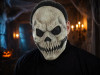 A person wearing a terrifying skull mask with black mesh eyes and sharp teeth, in a dimly lit room decorated with cobwebs and candles, with a jack-o-lantern visible in the background.
