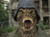 A close-up of a person wearing a grotesque monster mask with sharp teeth and a yellow eye, dressed in a military helmet and jacket. The background shows a dilapidated building and overgrown foliage.