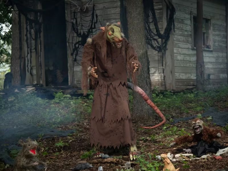 A creepy, tall rat-like creature stands in front of a dilapidated house surrounded by smaller rat figures, a monkey, and an alligator decoration, creating a Halloween scene.