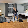 A woman is vacuuming the hardwood floor of a hotel room. A Nilfisk canister vacuum sits behind her while she cleans near the bed and a seating area. The room includes a window with curtains, modern furniture, and soft ambient lighting.