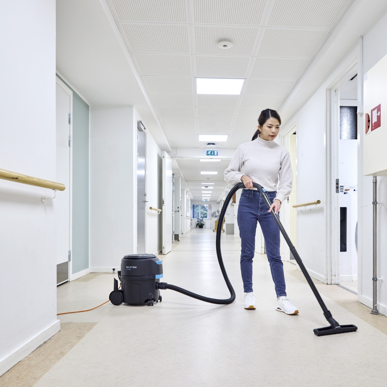 A woman is vacuuming the floor of a clean, white hospital or healthcare hallway using a Nilfisk canister vacuum positioned behind her. The corridor has safety rails along the walls, bright ceiling lights, and medical rooms on both sides.