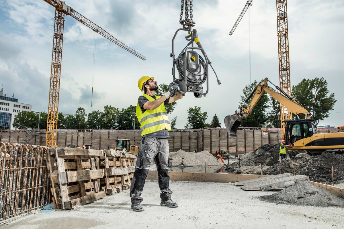 Operator using a Karcher cage-frame pressure washer to clean heavy equipment tracks on a muddy construction jobsite.