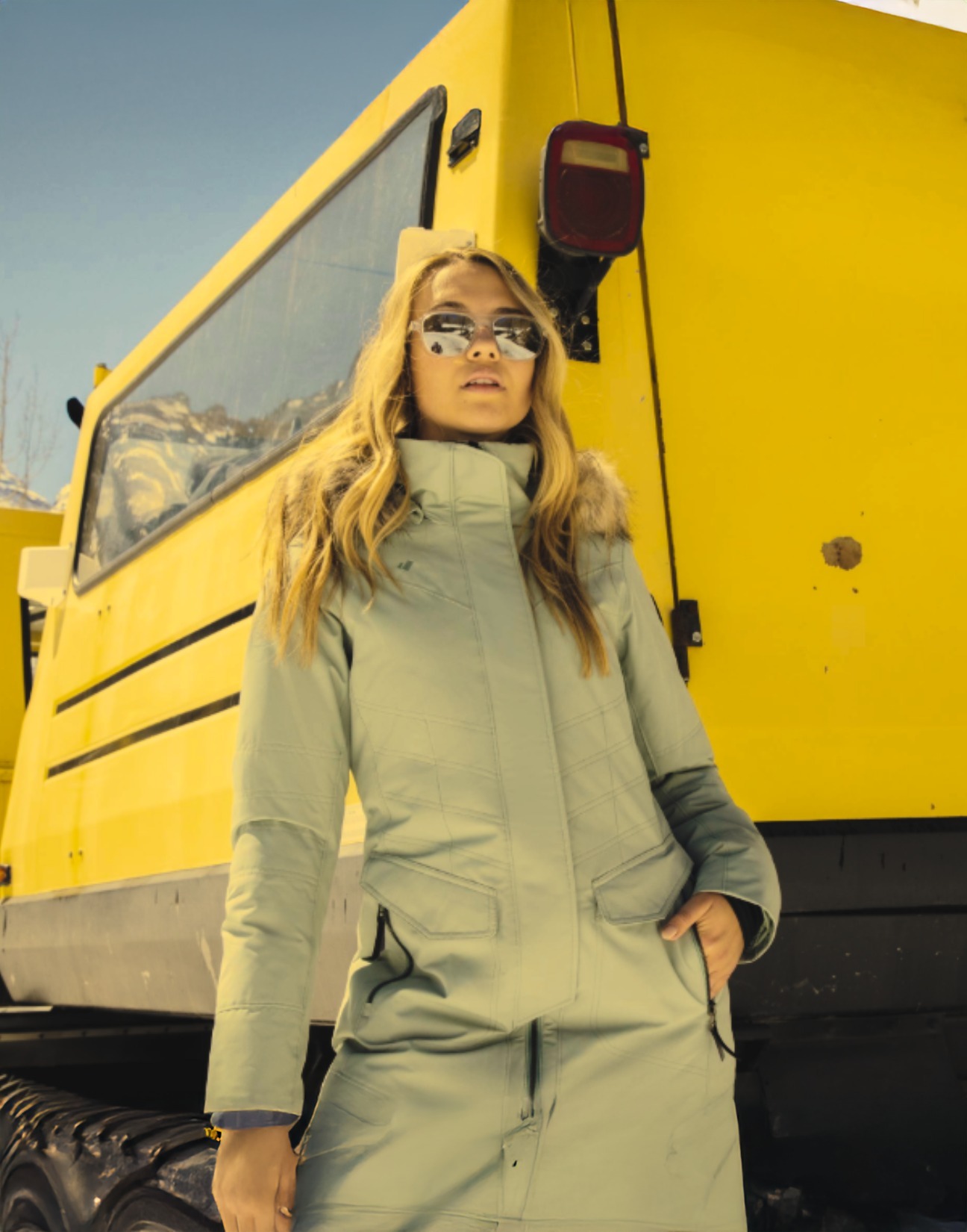 A woman standing in front of a yellow snow cat in her Obermeyer ski jacket and pants.