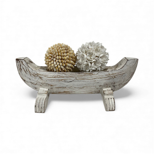 A whitewashed wooden bowl resembling a fishing boat, with intricate carvings, adds a coastal vibe and rustic charm to any room's decor as a unique centerpiece.