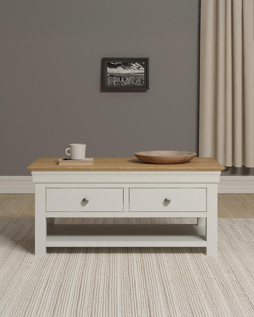 Vermont 2 Drawer Coffee Table, ivory and oak finish with a wooden top, two drawers with metallic knobs, placed in a living room setting with a gray wall, a small picture frame, a mug, and a wooden bowl on top.
