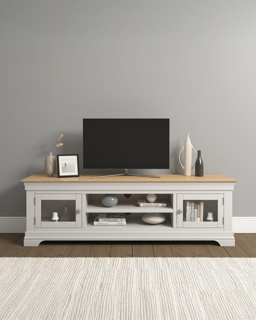 Vermont TV Stand in Ivory/Oak VM300-305-062, modern white wooden TV stand with glass-front cabinets and a light oak top surface, decorated with vases, a picture frame, and books, placed against a plain wall in a living room setting Vermont TV Stand in Ivory/Oak VM300-305-062, modern white wooden TV stand with glass-front cabinets and a light oak top surface, decorated with vases, a picture frame, and books, placed against a plain wall in a living room setting