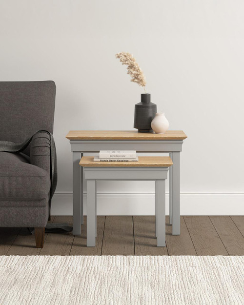Vermont Nest of 2 Tables in Pebble Grey/Oak VM300-303-043, a set of two nesting tables with light grey painted legs and natural oak tops, placed beside a grey armchair with decorative vases and books on top against a white wall.