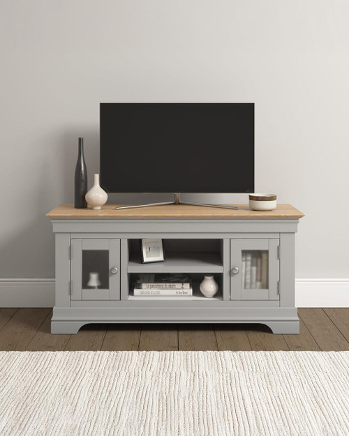 Vermont Small TV Stand in Pebble Grey/Oak VM300-305-063, modern TV stand with cabinet doors, open shelf, and decorative vases, placed in a living room with a flat-screen TV on top. Vermont Small TV Stand in Pebble Grey/Oak VM300-305-063, modern TV stand with cabinet doors, open shelf, and decorative vases, placed in a living room with a flat-screen TV on top.