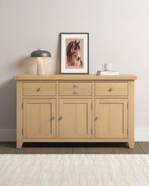 Hampton Oak 3 Door 3 Drawer Sideboard HO400-405-049, a wooden sideboard with a natural oak finish featuring three doors and three drawers, situated against a white wall with decorative items on top. Hampton Oak 3 Door 3 Drawer Sideboard HO400-405-049, a wooden sideboard with a natural oak finish featuring three doors and three drawers, situated against a white wall with decorative items on top.