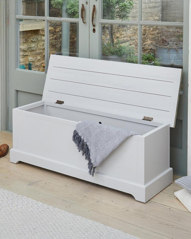 Signature Grey Hallway Storage Bench CFF20B, white wooden storage bench with a lift-up lid and a gray throw blanket draped over it, placed in a sunlit room with glass doors and a wooden floor.