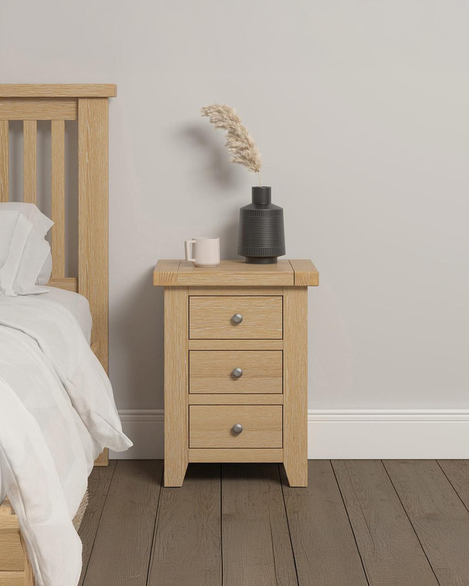 Hampton Oak 3 Drawer Bedside HO100-103-116, a wooden bedside table with three drawers and metallic knobs, placed next to a bed with light-colored bedding, decorated with a dark gray vase and a white mug on top, in a modern bedroom setting.