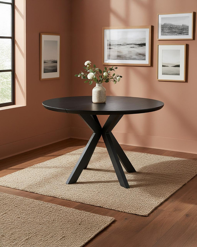 Hamilton Black Ash Round 120cm - 160cm Extending Table, black wooden round dining table with crisscross legs in a cozy room with pink walls and framed artwork.